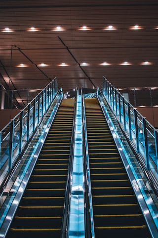 Twin escalators ascend in parallel under a ceiling with evenly spaced lights. The escalators are enclosed in glass with metal railings and have a sleek, modern appearance. The lighting creates a reflective effect on the shiny surfaces.