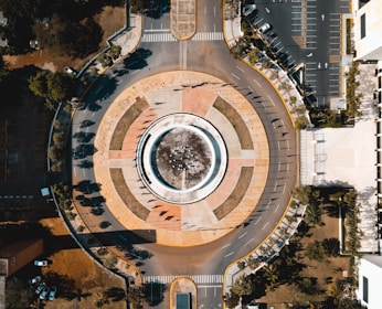 An aerial view of a large circular roundabout surrounded by roads, parking lots, and greenery. The center of the roundabout features a fountain or structure with pathways radiating outward. Shadows from nearby trees and buildings are visible, casting intricate patterns across the area.