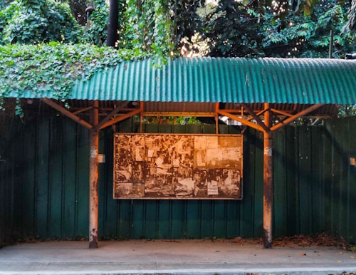 A green corrugated metal roof covers an outdoor bulletin board surrounded by dense foliage. The board is weathered and plastered with various papers and notices. The setting appears to be natural with lush greenery and a peaceful atmosphere.