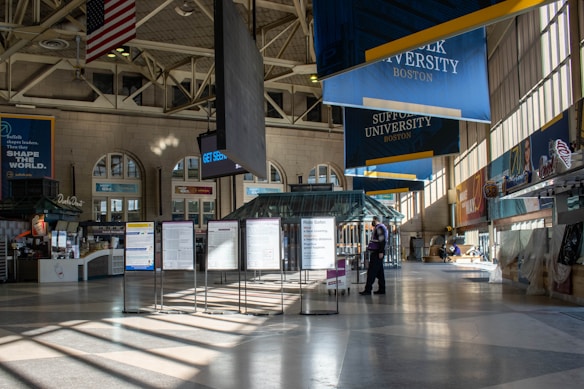 A spacious indoor transit terminal with high ceilings and large windows allowing natural light to stream in. There are various signs, including one for Suffolk University and directional boards. A few people are seen walking or standing, and there's an American flag hanging from the ceiling.