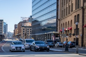A busy urban street scene with multiple cars at an intersection. Large buildings with glass and brick facades line the street, and a 'Boston Wharf Industrial' sign is visible. Red awnings and various signs are present on the right side, and the street is bustling with traffic.