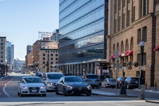 A busy urban street scene with multiple cars at an intersection. Large buildings with glass and brick facades line the street, and a 'Boston Wharf Industrial' sign is visible. Red awnings and various signs are present on the right side, and the street is bustling with traffic.