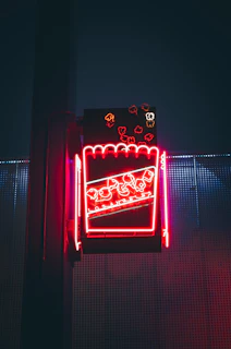 A neon sign depicting a popcorn box mounted on a structure, glowing with red and pink hues against a dark blue background. The sign features stylized popcorn pieces scattered around the box, creating a vibrant and eye-catching display.