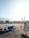 An airport tarmac with luggage carts and a small vehicle parked on the ground. A person wearing a safety vest walks near the carts. In the background, there's a view of distant mountains and palm trees. The scene is bathed in warm sunlight, creating long shadows.