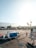 An airport tarmac with luggage carts and a small vehicle parked on the ground. A person wearing a safety vest walks near the carts. In the background, there's a view of distant mountains and palm trees. The scene is bathed in warm sunlight, creating long shadows.