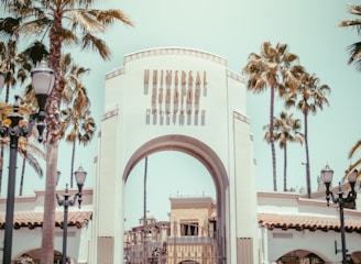 An iconic archway entrance with the words 'Universal Studios Hollywood' written on it, surrounded by tall palm trees. The scene is set on a sunny day with a clear sky. Decorative street lamps frame the entrance, adding to the theme park ambiance.