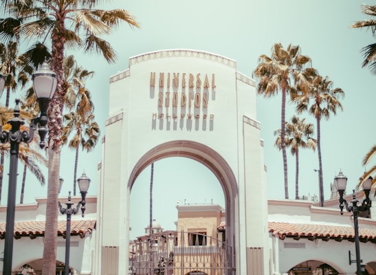 An iconic archway entrance with the words 'Universal Studios Hollywood' written on it, surrounded by tall palm trees. The scene is set on a sunny day with a clear sky. Decorative street lamps frame the entrance, adding to the theme park ambiance.