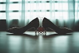 Two decorative high-heeled shoes are positioned in a mirrored arrangement against a softly lit curtain backdrop. The shoes feature intricate detailing and are connected by a decorative red symbol with heart-shaped cutouts.