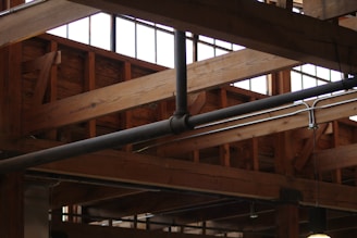 Wooden beams and metal pipes are visible on the ceiling of an industrial-style building. Large windows provide natural light, highlighting the structure's rustic and robust design.