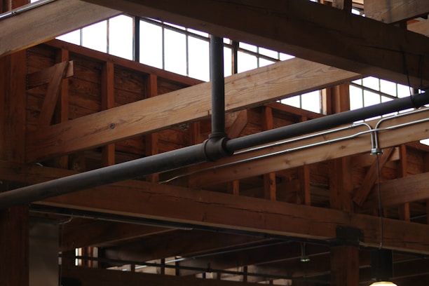 Wooden beams and metal pipes are visible on the ceiling of an industrial-style building. Large windows provide natural light, highlighting the structure's rustic and robust design.