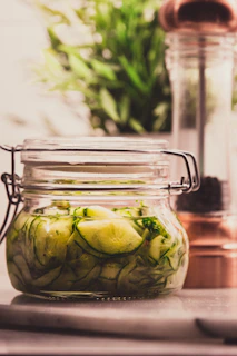 A jar of homemade lacto-fermented cucumbers with fresh herbs beside it on a minimalist kitchen counter.