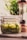 A glass jar containing sliced cucumbers and herbs, placed on a countertop. There is a blurred background with a green plant and a pepper mill beside the jar.