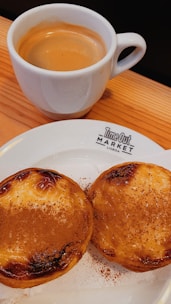 A cup of espresso is placed on a wooden surface next to a plate containing two Portuguese custard tarts sprinkled with cinnamon. The plate has 'Time Out Market Lisboa' printed on it.