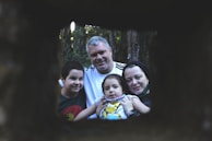 A family of four poses for a portrait outdoors, framed by a dark, rectangular opening. The setting is within a forest, with trees visible in the background. The family looks content and close-knit, with the parents and two children all focusing on the camera.