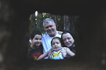 A family of four poses for a portrait outdoors, framed by a dark, rectangular opening. The setting is within a forest, with trees visible in the background. The family looks content and close-knit, with the parents and two children all focusing on the camera.