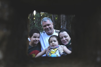 A family of four poses for a portrait outdoors, framed by a dark, rectangular opening. The setting is within a forest, with trees visible in the background. The family looks content and close-knit, with the parents and two children all focusing on the camera.