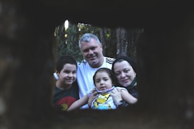 A family of four poses for a portrait outdoors, framed by a dark, rectangular opening. The setting is within a forest, with trees visible in the background. The family looks content and close-knit, with the parents and two children all focusing on the camera.