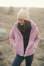 A person wearing a cozy pink fluffy jacket and a cream knit hat with a pom-pom, standing outdoors in a natural setting with a blurred background.