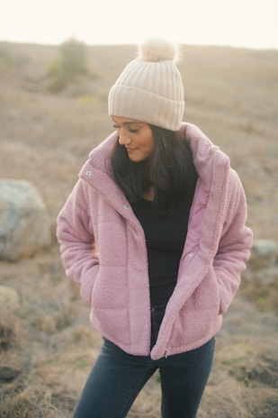 A person wearing a cozy pink fluffy jacket and a cream knit hat with a pom-pom, standing outdoors in a natural setting with a blurred background.