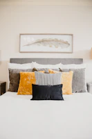 A well-made bed topped with multiple pillows of varying textures and colors, including a black pillow in the front, a black and white patterned pillow, and larger velvet pillows in gold and grey. Above the bed hangs a rectangular framed abstract artwork featuring a delicate leaf motif in muted tones.