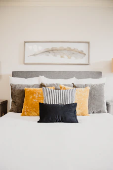 A well-made bed topped with multiple pillows of varying textures and colors, including a black pillow in the front, a black and white patterned pillow, and larger velvet pillows in gold and grey. Above the bed hangs a rectangular framed abstract artwork featuring a delicate leaf motif in muted tones.