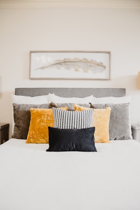 A well-made bed topped with multiple pillows of varying textures and colors, including a black pillow in the front, a black and white patterned pillow, and larger velvet pillows in gold and grey. Above the bed hangs a rectangular framed abstract artwork featuring a delicate leaf motif in muted tones.