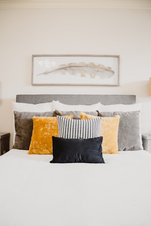 A well-made bed topped with multiple pillows of varying textures and colors, including a black pillow in the front, a black and white patterned pillow, and larger velvet pillows in gold and grey. Above the bed hangs a rectangular framed abstract artwork featuring a delicate leaf motif in muted tones.