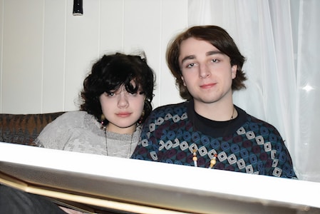 A young woman and man sit closely on a couch. They are wearing casual clothing and necklaces. The background features white curtains and a soft-lit indoor setting.