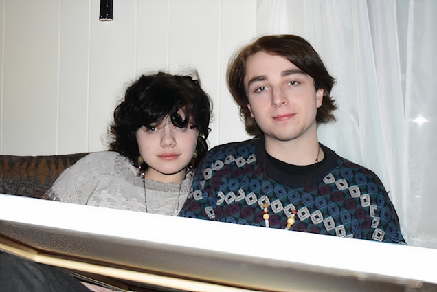 A young woman and man sit closely on a couch. They are wearing casual clothing and necklaces. The background features white curtains and a soft-lit indoor setting.