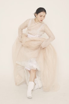 A woman is dressed in a traditional, flowing garment with soft, translucent layers. Her attire includes a long, beige dress with white underlayers and matching boots. She is captured in a graceful pose, looking downwards with her hands gathered near her waist. Her hair is neatly styled and pinned back, complementing the elegantly serene setting.