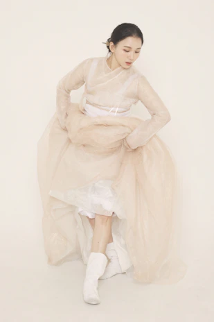 A woman is dressed in a traditional, flowing garment with soft, translucent layers. Her attire includes a long, beige dress with white underlayers and matching boots. She is captured in a graceful pose, looking downwards with her hands gathered near her waist. Her hair is neatly styled and pinned back, complementing the elegantly serene setting.