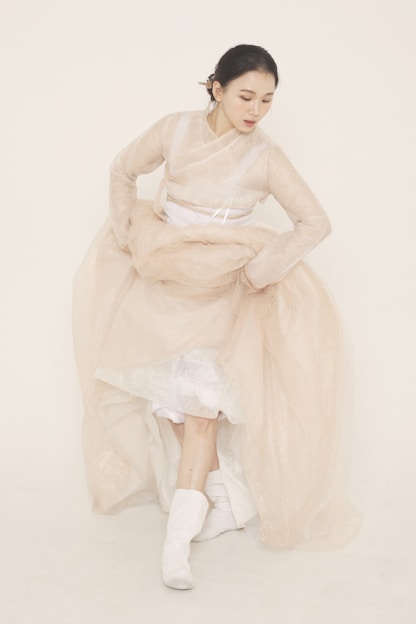 A graceful woman wearing a flowing modest dress in soft earth tones, standing in a sunlit urban courtyard.