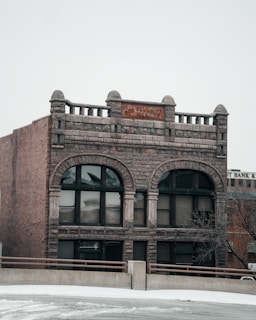 A historic-looking stone building with arched windows and an inscription reading 'Odd Fellows Hall' above the entrance. The architecture includes decorative stonework and a wrought iron balcony. The building appears weathered, adding to its vintage charm. Snow covers the ground in front of the structure.