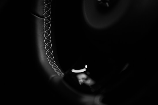 Close-up view of a car's steering wheel, showcasing intricate stitching along the edge. The image features a black and white color scheme, emphasizing the texture and craftsmanship.