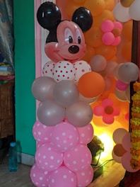 A decorative balloon arrangement resembling a cartoon character with a large head featuring black ears and a smiling face. The balloons are predominantly pink with white polka dots, accompanied by others in pastel shades. The background includes more balloons in orange, pink, and white colors.