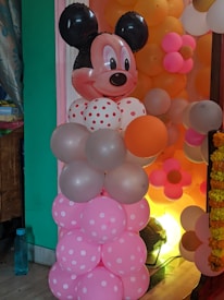 A decorative balloon arrangement resembling a cartoon character with a large head featuring black ears and a smiling face. The balloons are predominantly pink with white polka dots, accompanied by others in pastel shades. The background includes more balloons in orange, pink, and white colors.