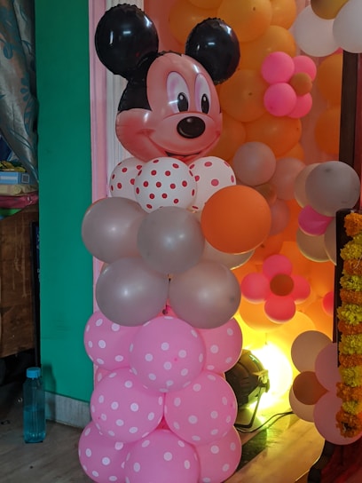 A decorative balloon arrangement resembling a cartoon character with a large head featuring black ears and a smiling face. The balloons are predominantly pink with white polka dots, accompanied by others in pastel shades. The background includes more balloons in orange, pink, and white colors.