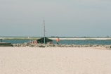 A sandy beach with a rocky shoreline and a tent-like structure near the water. Two people in orange clothing are standing near the tent. In the background, there are several small islands and a calm blue sea.