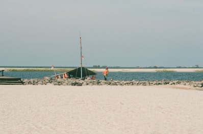 A sandy beach with a rocky shoreline and a tent-like structure near the water. Two people in orange clothing are standing near the tent. In the background, there are several small islands and a calm blue sea.