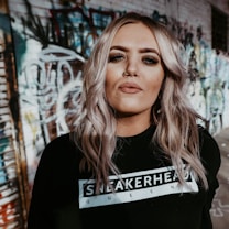 A person with long, wavy blonde hair stands in front of a graffiti-covered wall. They are wearing a black sweatshirt with the text 'SNEAKERHEAD QUEENS' and have facial piercings. The image has an urban, edgy vibe.