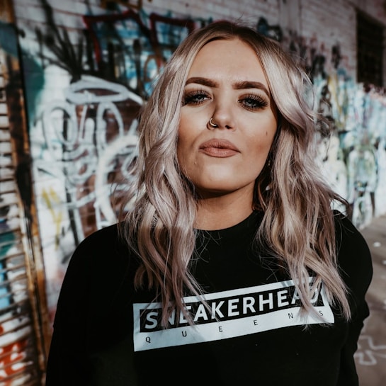 A person with long, wavy blonde hair stands in front of a graffiti-covered wall. They are wearing a black sweatshirt with the text 'SNEAKERHEAD QUEENS' and have facial piercings. The image has an urban, edgy vibe.