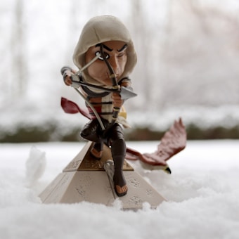 A figurine of an archer wearing a hooded outfit, poised on a small pyramid with a bow and arrow drawn, surrounded by snow. A falcon is flying alongside the figure, suggesting movement.