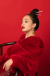 A person is elegantly posing against a vibrant red background. They are wearing a luxurious red fur coat and have a distinctive hairstyle with decorative chopsticks. Their makeup is bold, featuring striking red lipstick that matches the overall color theme.