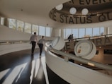 A modern interior space with a sweeping curved ramp, showcasing a couple walking hand in hand. The architecture features large circular texts and round light fixtures on the ceiling. Windows reveal a glimpse of autumn trees outside, with warm sunlight casting long shadows on the floor.