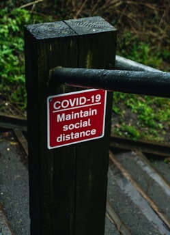 A red sign with white text that reads 'COVID-19 Maintain social distance' is mounted on a dark wooden post. The background shows an outdoor setting with green foliage and a partially visible path or stairway, suggesting a natural environment.