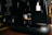 A close-up view of an espresso machine dispensing coffee into a glass, with a small sculpture and a framed quote in the background. The countertop is wooden, and the lighting is dim, creating a cozy atmosphere.