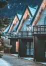 A row of wooden cabins adorned with string lights along the eaves, set against a dark forest backdrop. The triangular roofs create a cozy, picturesque scene with warm light glowing from the windows, contrasting with the cold, snowy surroundings.