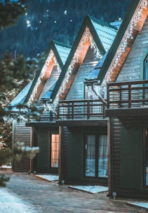 A row of wooden cabins adorned with string lights along the eaves, set against a dark forest backdrop. The triangular roofs create a cozy, picturesque scene with warm light glowing from the windows, contrasting with the cold, snowy surroundings.