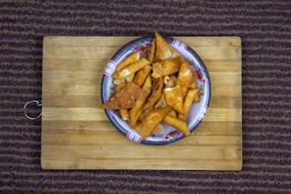 A plate filled with golden-brown, crispy triangular snacks is placed on a wooden cutting board. The plate has a floral pattern on the edge and is situated atop a carpet with a striped pattern.