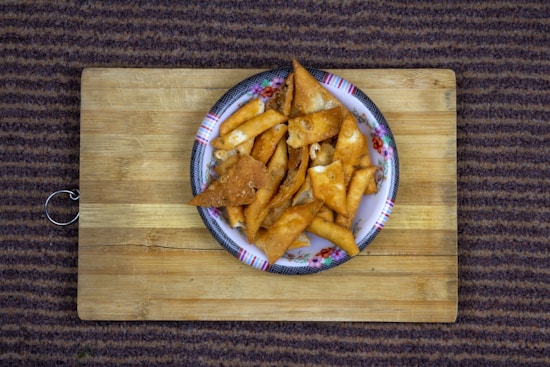 A plate filled with golden-brown, crispy triangular snacks is placed on a wooden cutting board. The plate has a floral pattern on the edge and is situated atop a carpet with a striped pattern.